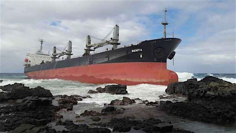 The cargo ship Benita is beached on a rocky shoreline with waves crashing against its hull under a cloudy sky.