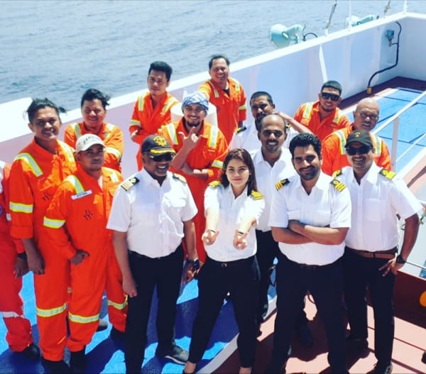 A smiling, diverse group of seafarers in orange coveralls and white officer uniforms poses for a photo on a ship deck with the ocean in the background.
