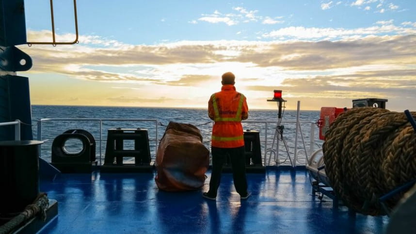 A male seafarer in a high-visibility jacket stands on the deck of a ship, looking out at the sun setting over the ocean.